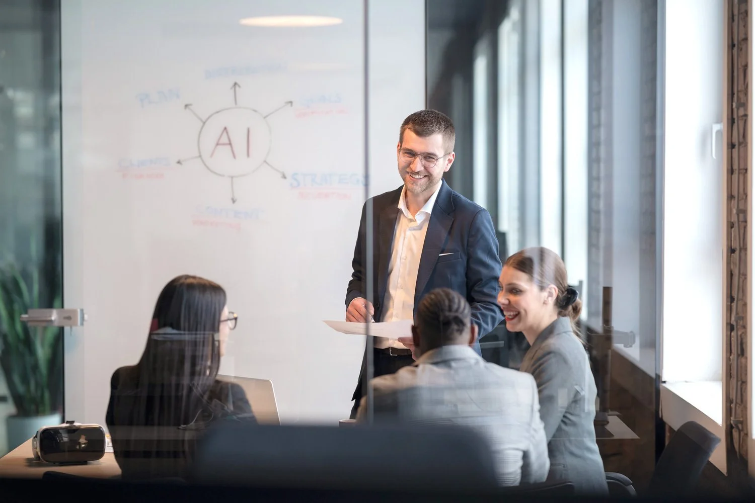 Geschäftsleute in einem Meeting, ein Mann steht mit Papier in der Hand und lächelt, während vier Frauen am Tisch sitzen und lachen, im Hintergrund eine Whiteboard mit 'AI' und Strategiethemen.