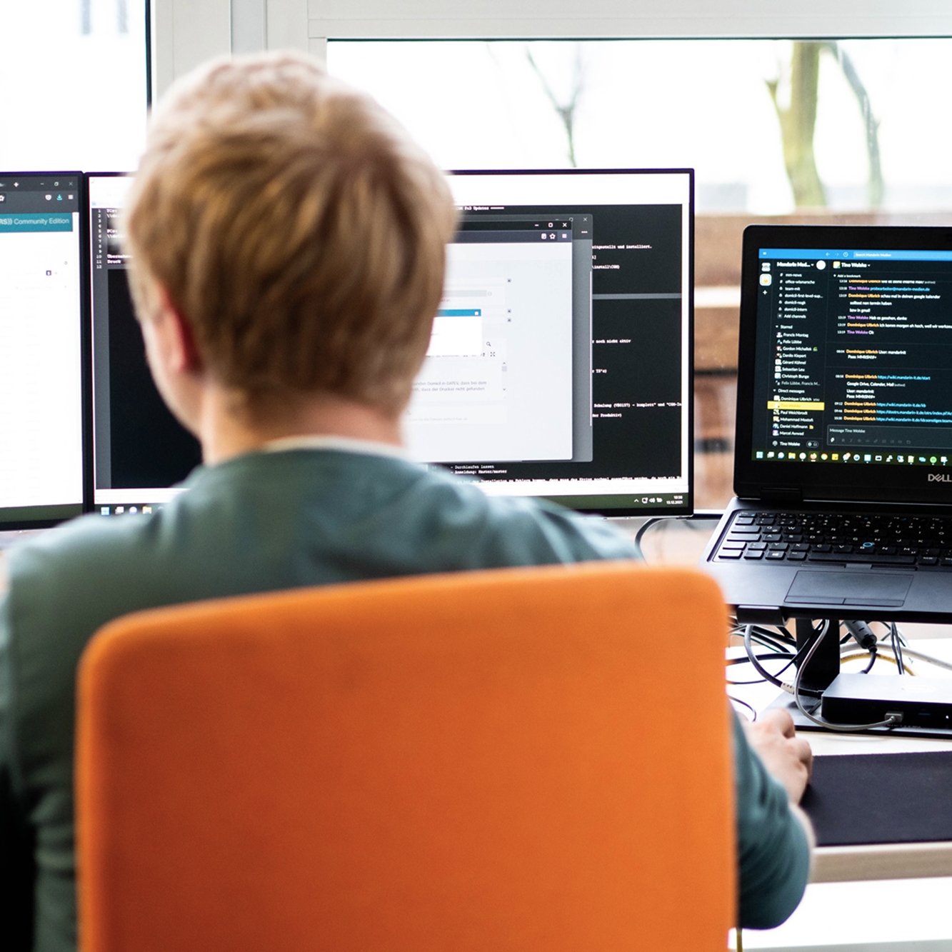 Person sitzt an einem Schreibtisch mit drei Monitoren, arbeitet an einem Computer, die Monitore zeigen Programmier- oder technischen Code auf dunklem Hintergrund.