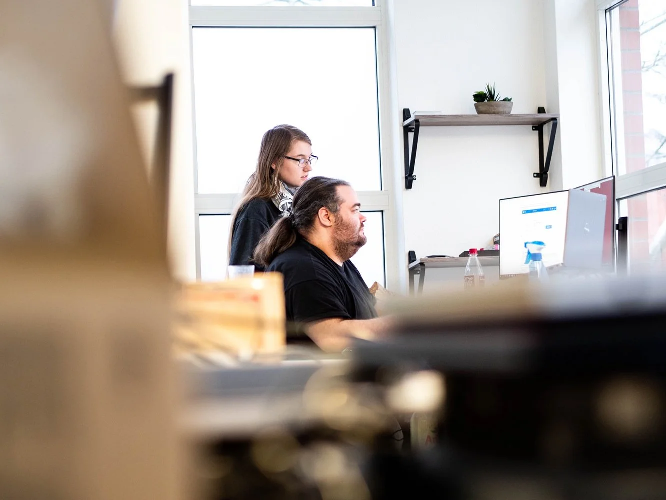Zwei Menschen, ein Mann mit langen Haaren und ein Bart, und eine Frau mit Brille, sitzen nebeneinander an Tischen in einem hellen Raum. Sie schauen auf Computerbildschirme und arbeiten.