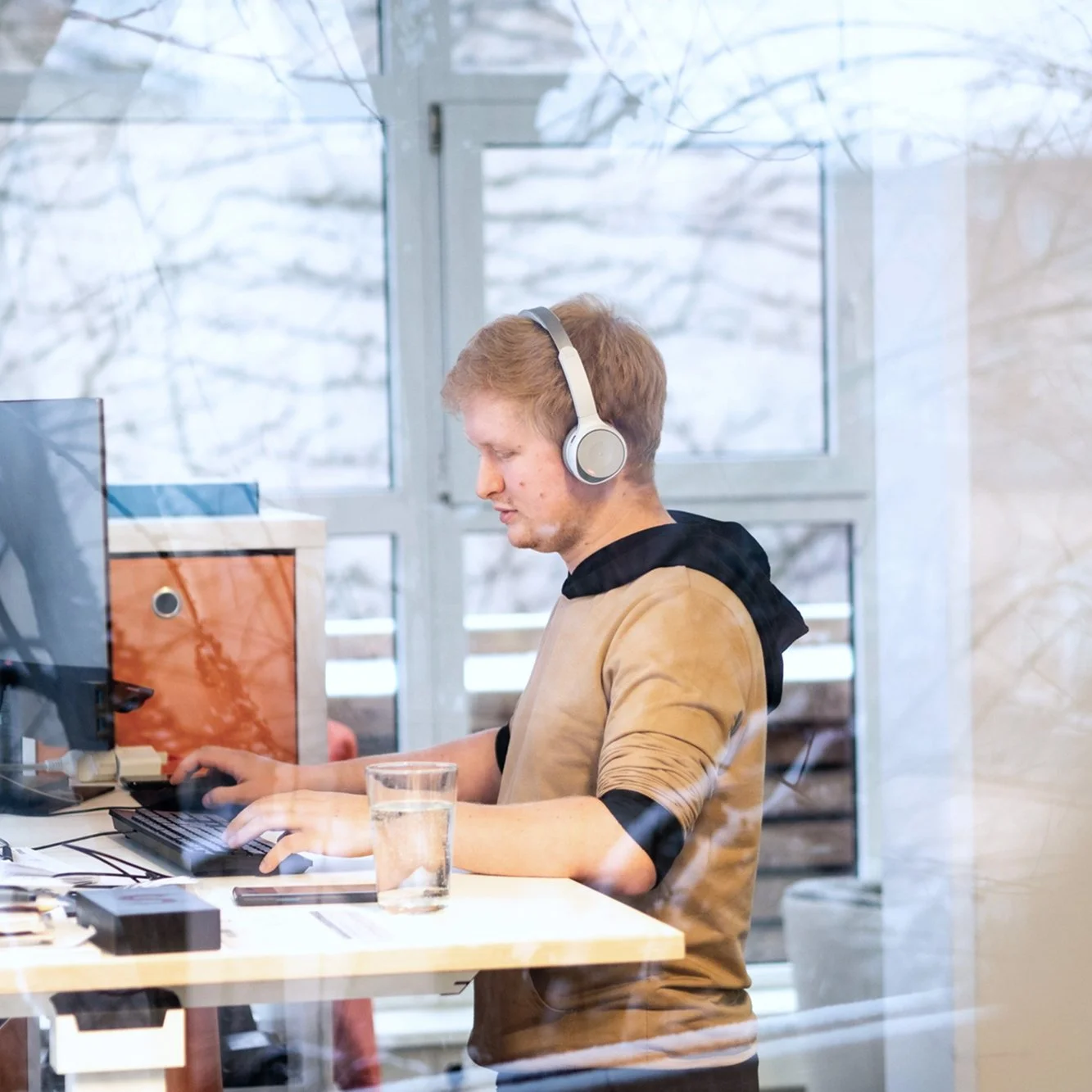 Junger Mann mit Kopfhörern arbeitet an einem Computer in einem Büro, sichtbar durch eine Glasscheibe, mit Winterlandschaft im Hintergrund.