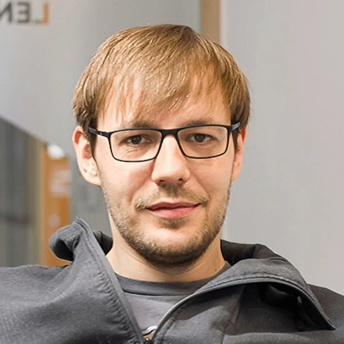 Ein junger Mann mit braunen Haaren, Bart und Brille, trägt eine graue Jacke und einen grauen Pullover, steht in einem Raum mit grauem Hintergrund.