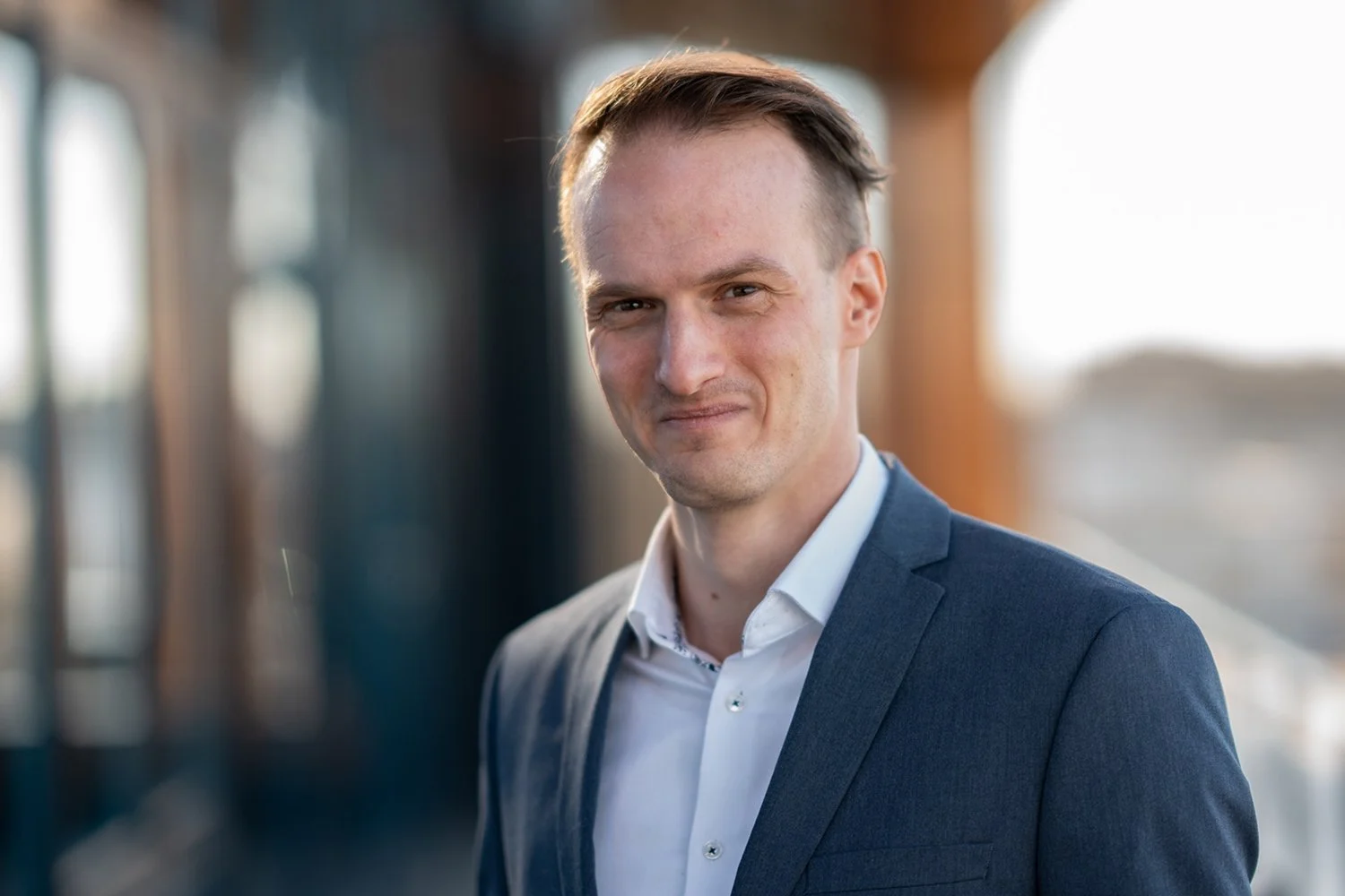 Junger Mann trägt einen dunklen Anzug und steht vor einem Gebäude mit großen Fenstern, im Sonnenlicht, lächelnd.