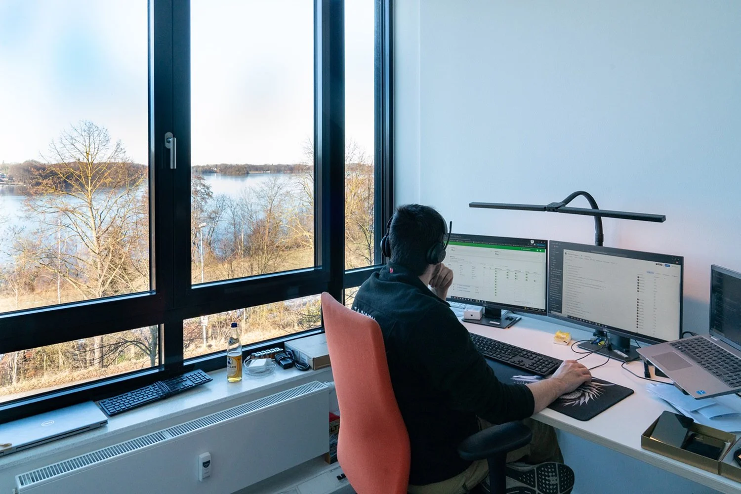 Mann arbeitet an einem Schreibtisch mit zwei Monitoren, tragt Kopfhörer und blickt nach draußen auf einen Fluss und Bäume bei Sonnenlicht.
