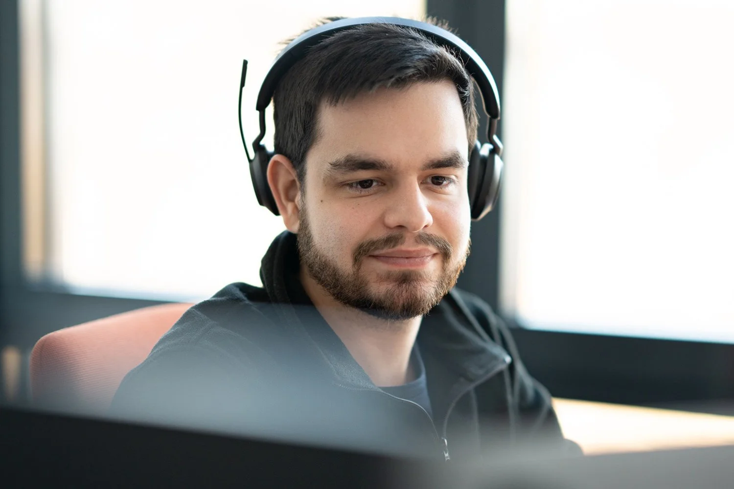 Ein junger Mann mit dunklem Haar und Bart trägt Kopfhörer und sitzt vor einem Computer, lächelnd und konzentriert, in einem hell beleuchteten Raum.