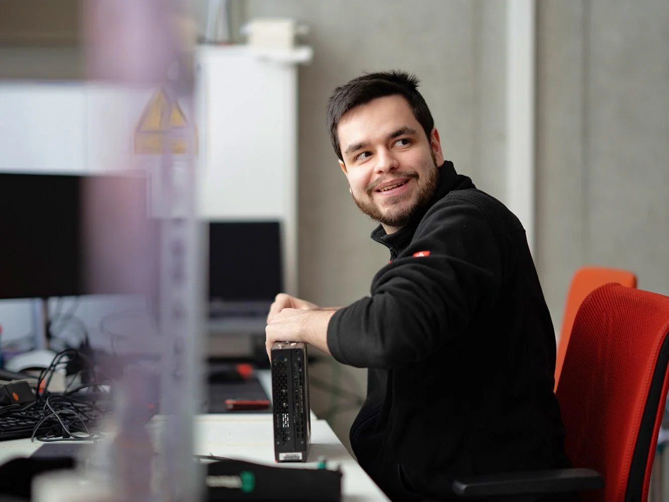 Junger Mann sitzt an einem Schreibtisch in einem Büro und lächelt, während er ein elektronisches Gerät hält.