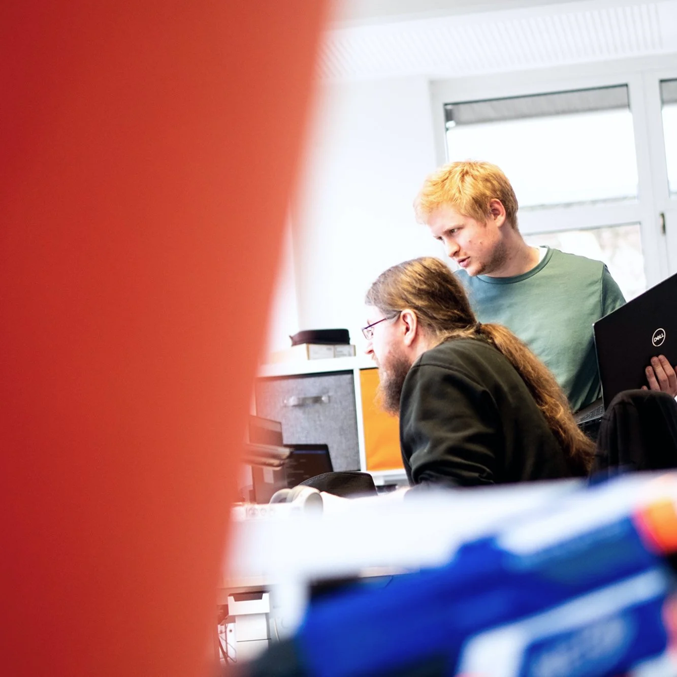 Zwei junge Männer mit Laptops in einem Büro, einer steht und der andere sitzt, während sie zusammenarbeiten.