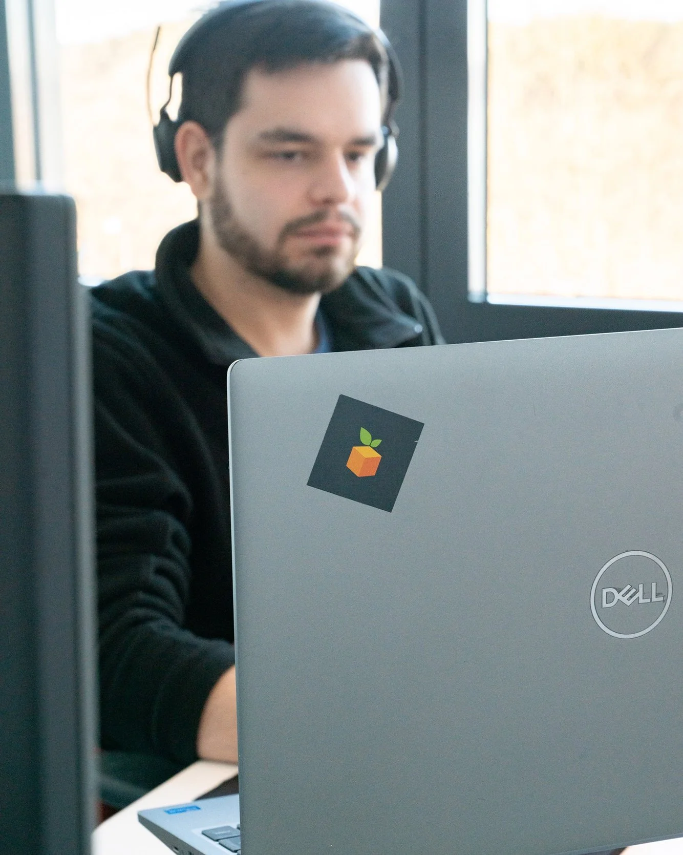 Junge Mann mit schwarzen Haaren und Bart, trägt Kopfhörer, sitzt an einem Laptop mit einem Sticker eines grünen Blattes und orange-braunem Würfel, in einem Raum mit Fenstern im Hintergrund.