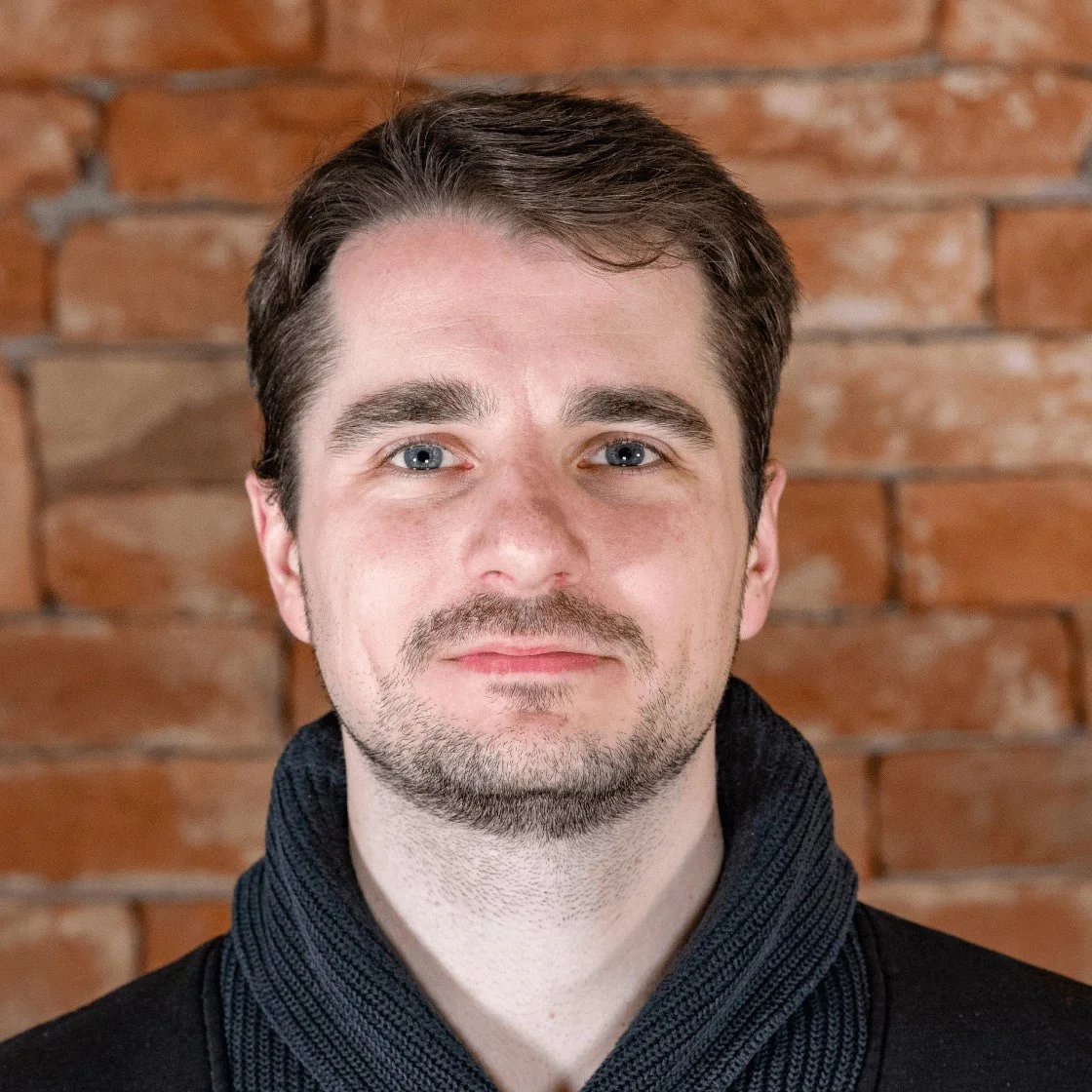 Nahaufnahme eines Mannes mit dunklem Haar und Bart vor einer Ziegelsteinwand.