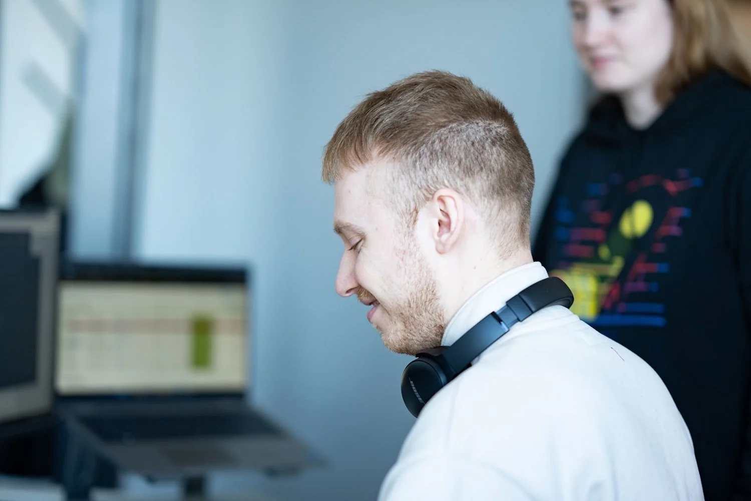 Ein junger Mann mit Kopfhörern sitzt vor einem Computerbildschirm, während eine Frau im Hintergrund neben ihm steht und zusieht.