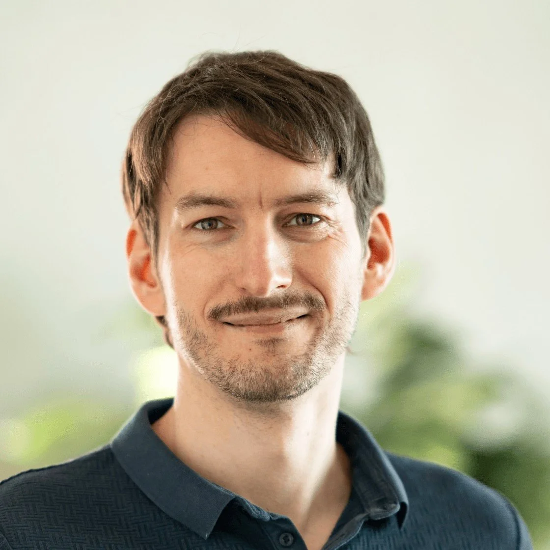 Porträt eines lächelnden Mannes mit dunklem Haar und Barthaars, trägt eine dunkle Hemd, vor unscharfen grünem Hintergrund.