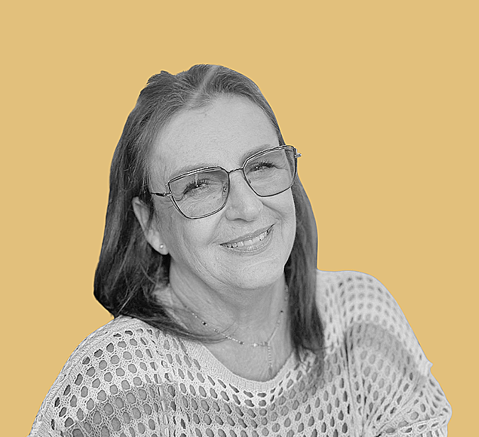 A smiling woman with glasses and shoulder-length hair, wearing a perforated light-colored top, sitting at a table with a glass of drink and a straw against a plain, mustard-colored background.