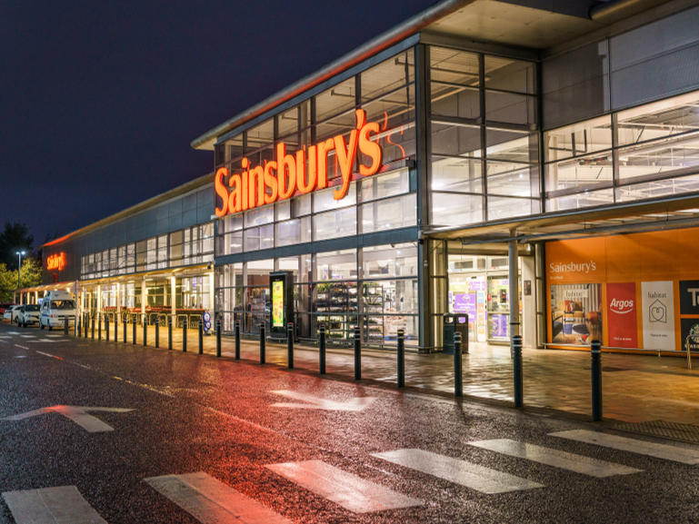Exterior view of a Sainsbury's supermarket at night with illuminated orange signage and large glass windows.