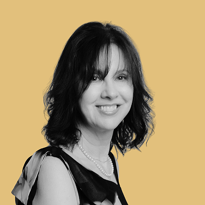 A woman with shoulder-length dark hair, smiling, wearing a black dress with a pearl necklace, against a beige background.