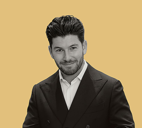 Black and white photo of a man in a suit, smiling in profile, holding a cleaning cloth against his face, against a solid light yellow background.