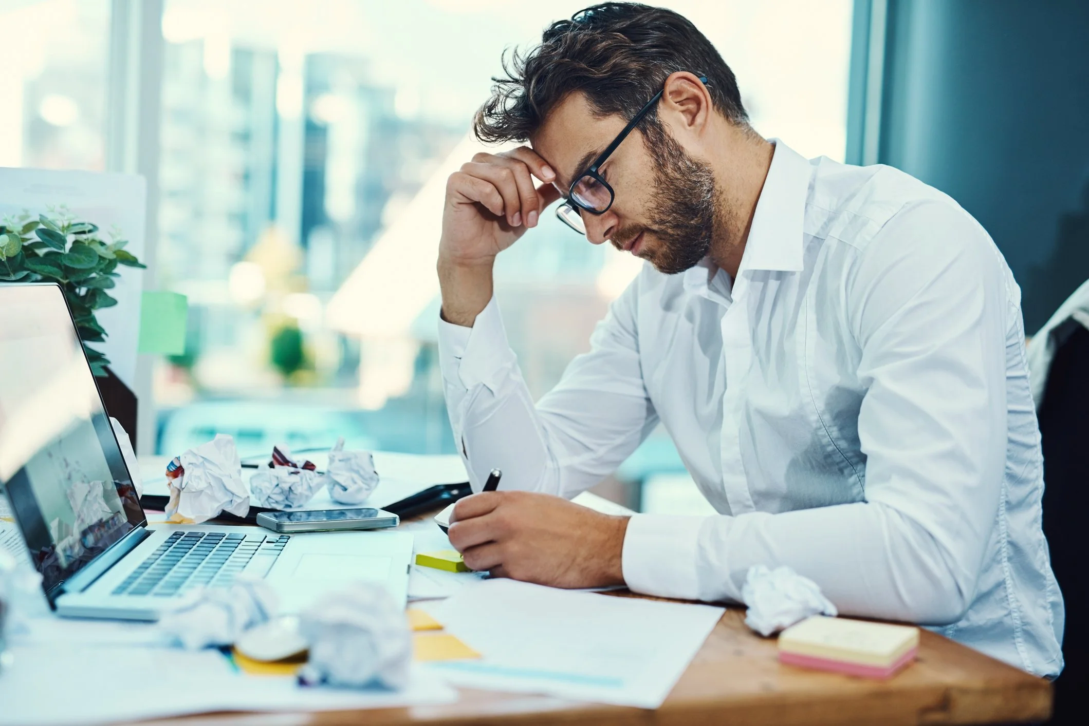 Frustrated man in white shirt sitting at cluttered desk in an office, holding forehead with one hand and writing with the other, surrounded by crumpled papers, sticky notes, a laptop, and a smartphone.