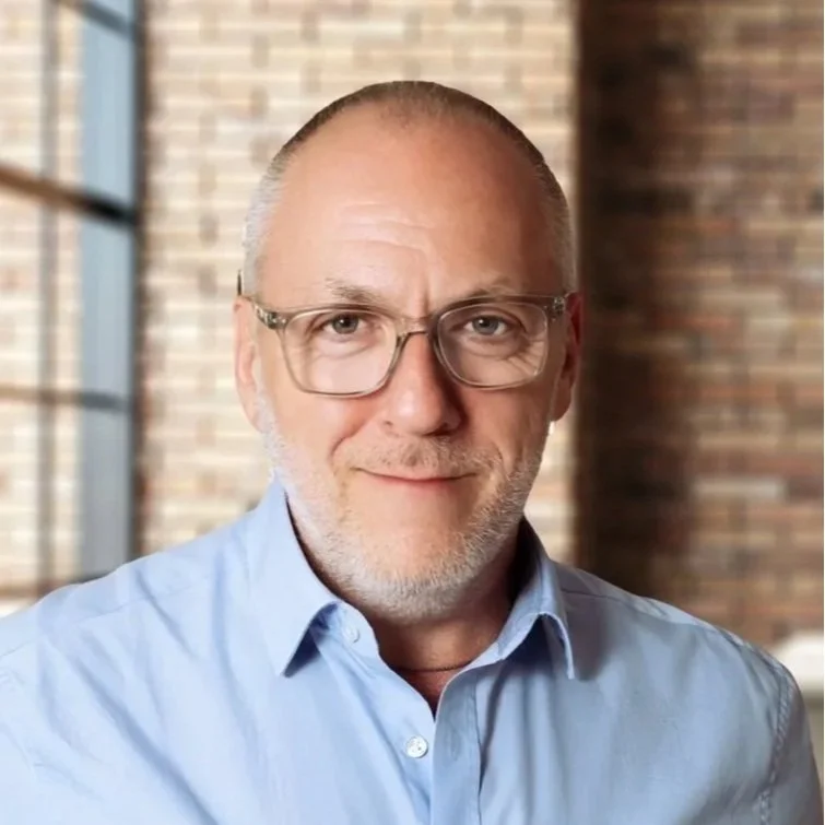 A middle-aged man with glasses, a short beard, wearing a light blue dress shirt, smiling in front of a blurred brick wall background.