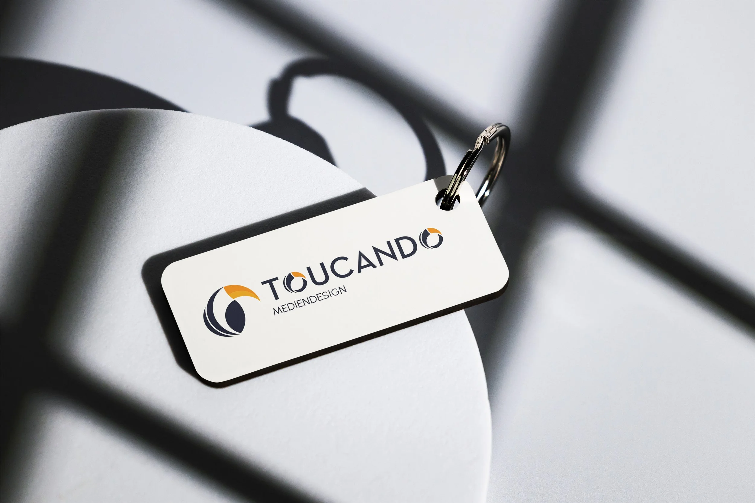 Namensschild mit Logo und Text auf einem weißen runden Objekt, Schatten sichtbar, an einem Metallhaken befestigt.