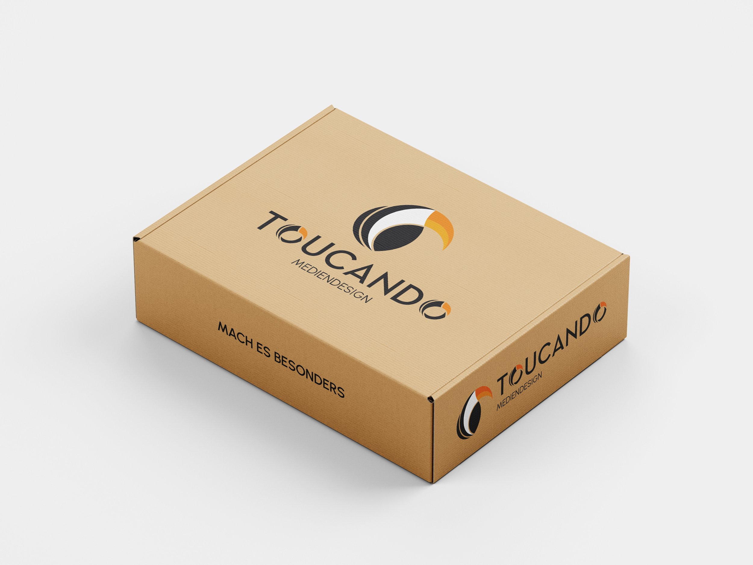 Ein brauner Karton mit Logo und Text. Das Logo besteht aus schwarzen, weißen und orangefarbenen Kurven, die eine Schildkröte andeuten. Der Text auf dem Karton lautet 'TOUCANDO MEDIENDESIGN' und 'MACH ES BESONDERS'.