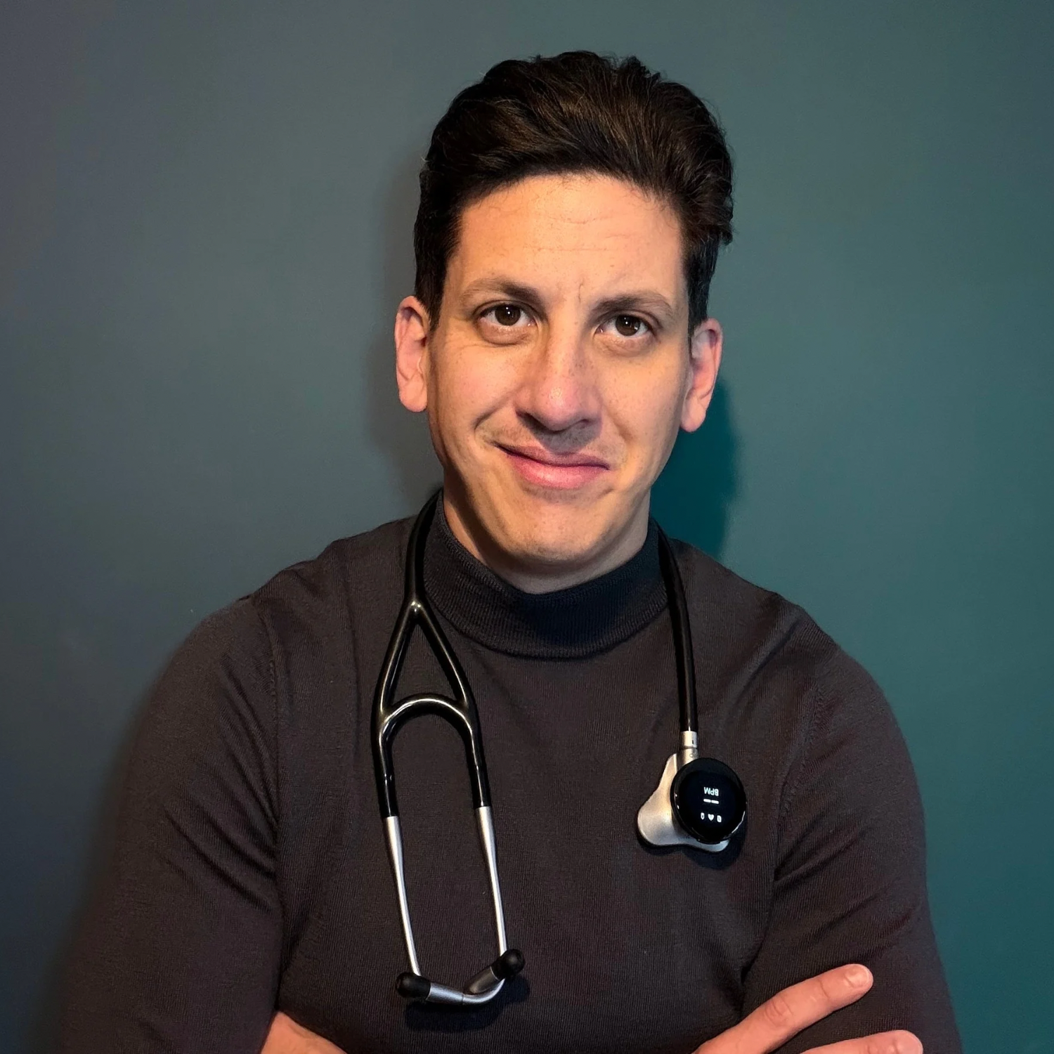 A male healthcare professional wearing a black shirt and a stethoscope around his neck, standing against a teal background.