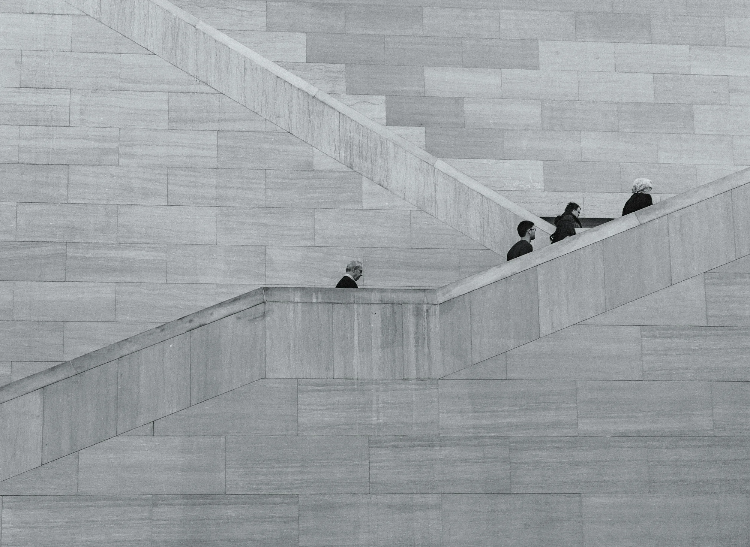 Black and white photo of people walking up a staircase with angular design and plain wall background.
