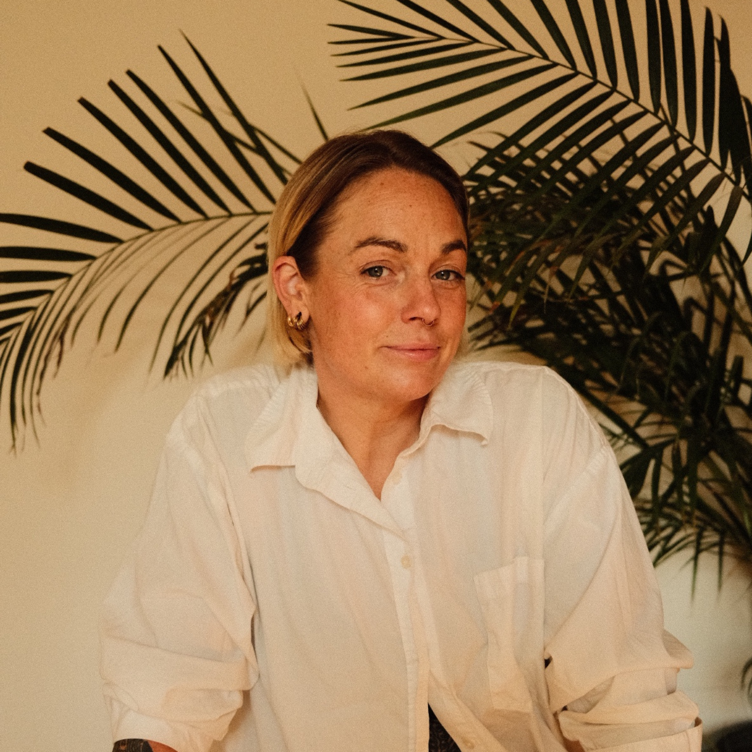 A woman with short blonde hair, wearing a white button-up shirt, sitting in front of a large green palm plant with a beige wall background.