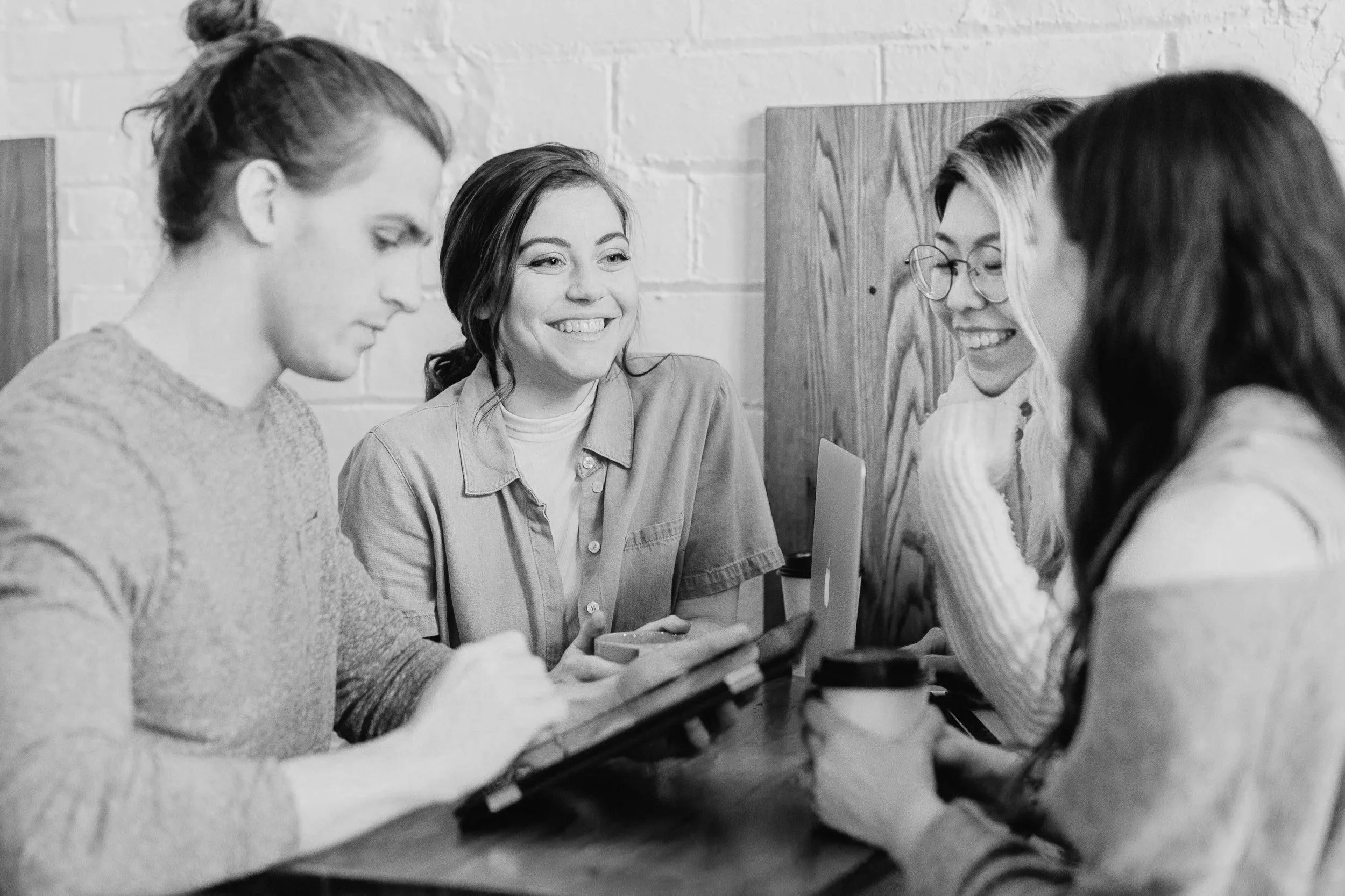 Four young women sitting together at a table, engaging with each other and devices, smiling and smiling in a casual setting.