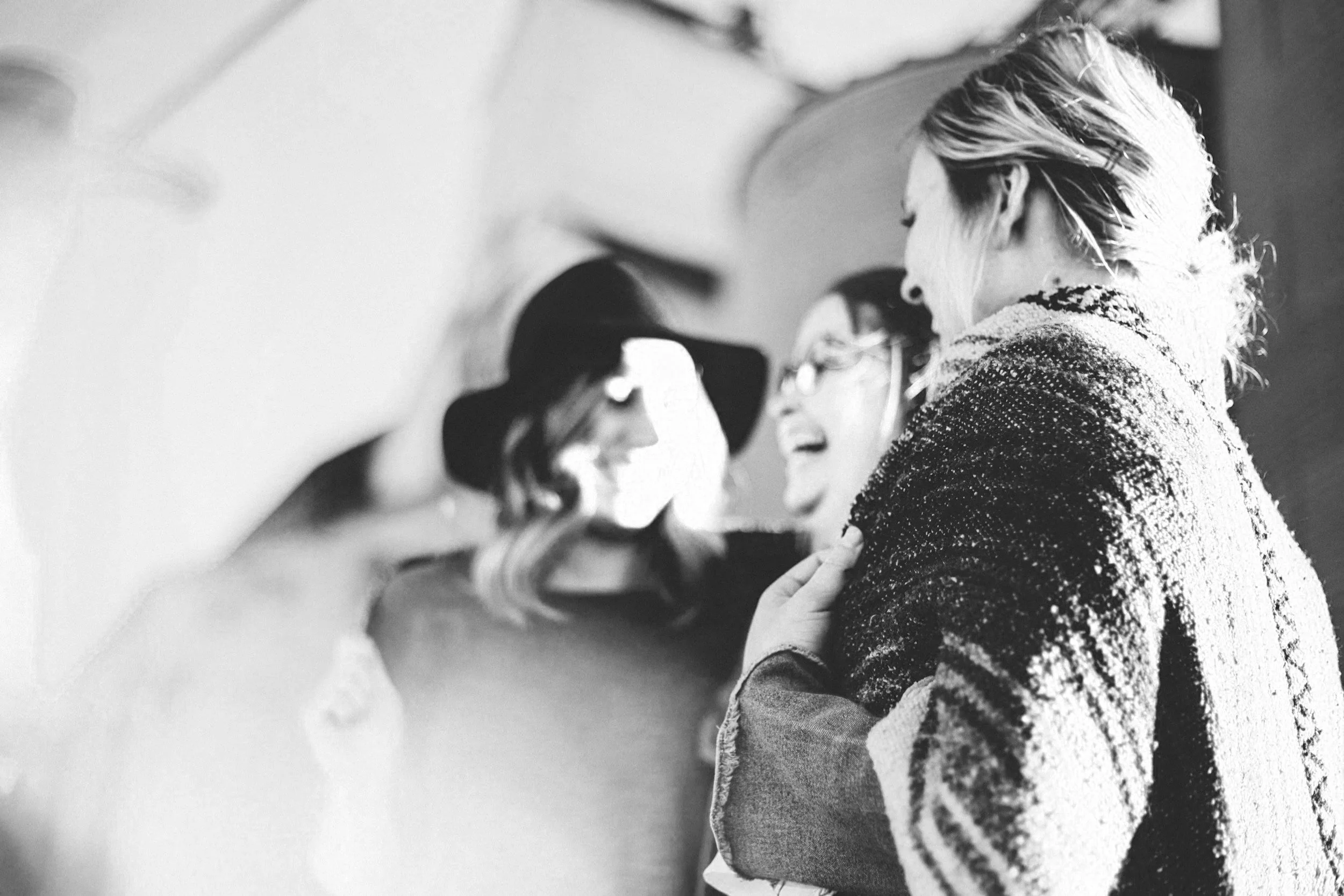 Three women laughing and smiling together, one wearing a hat and glasses, in black and white photograph.
