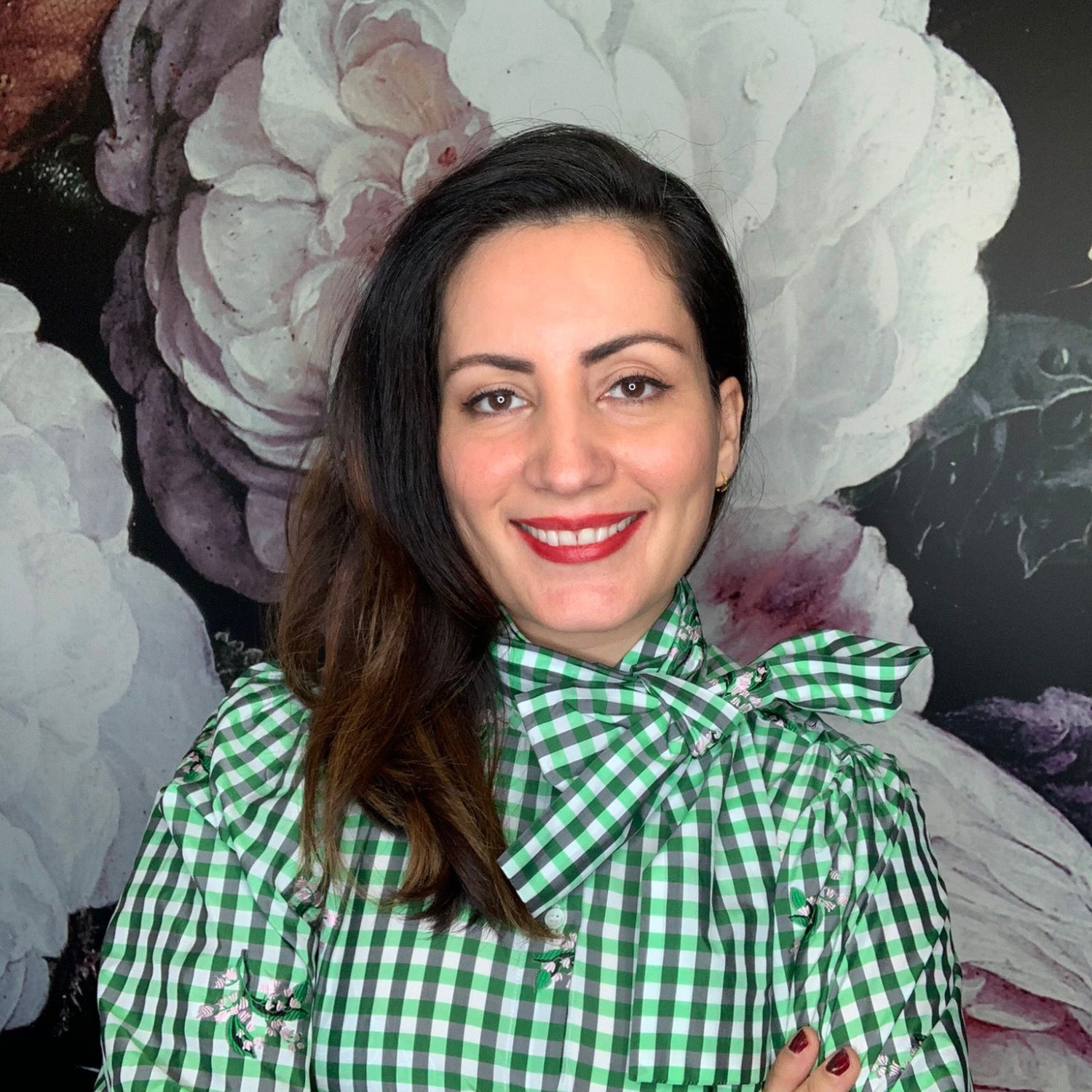 A woman with long, dark brown hair and light skin, wearing a green and white checkered blouse with a bow tie, stands in front of a floral mural background.