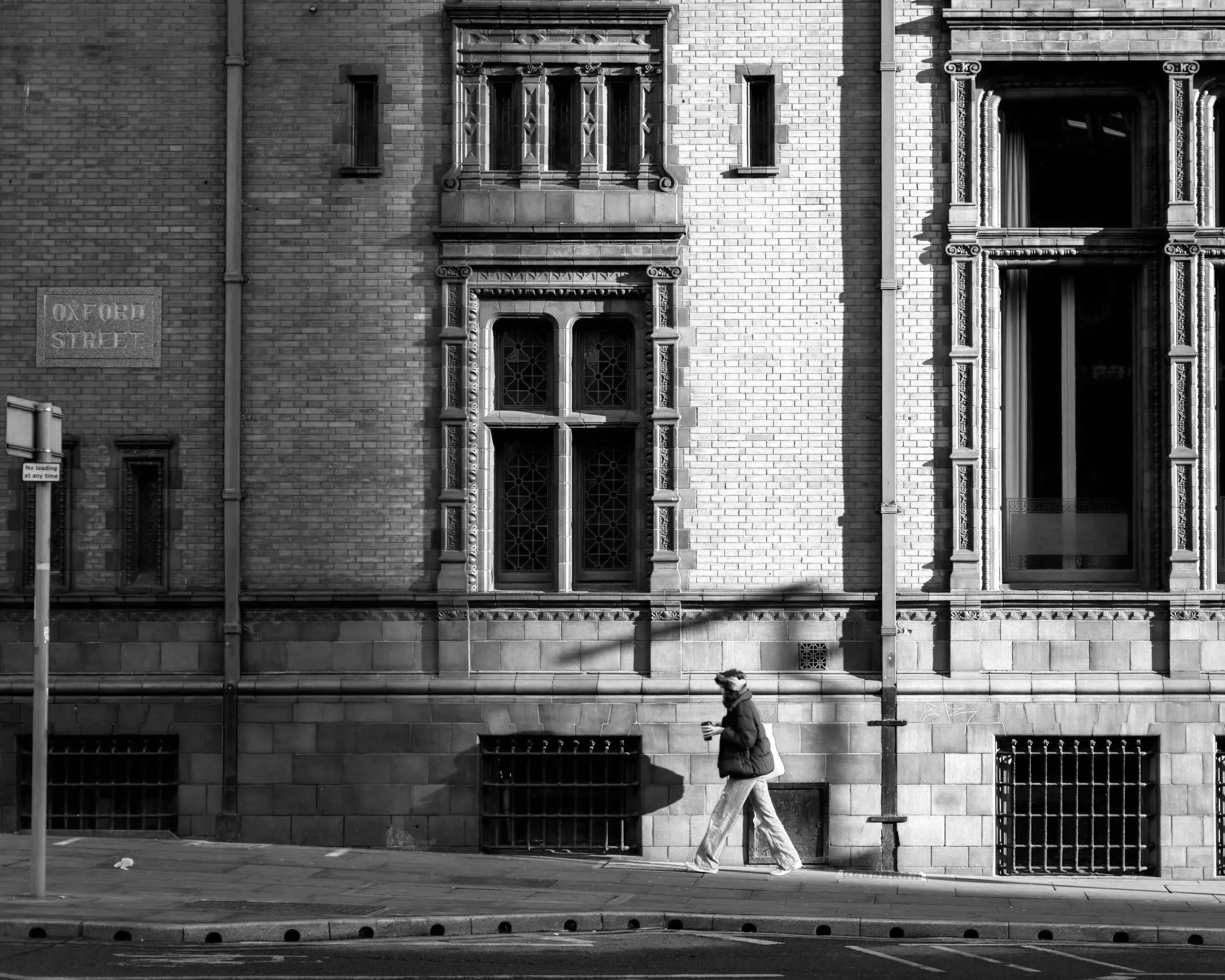A person wearing a jacket, pants, and a hat walking on a sidewalk with a beverage in hand, in front of a brick building with ornate windows and a street sign that says 'Oxford Street.'