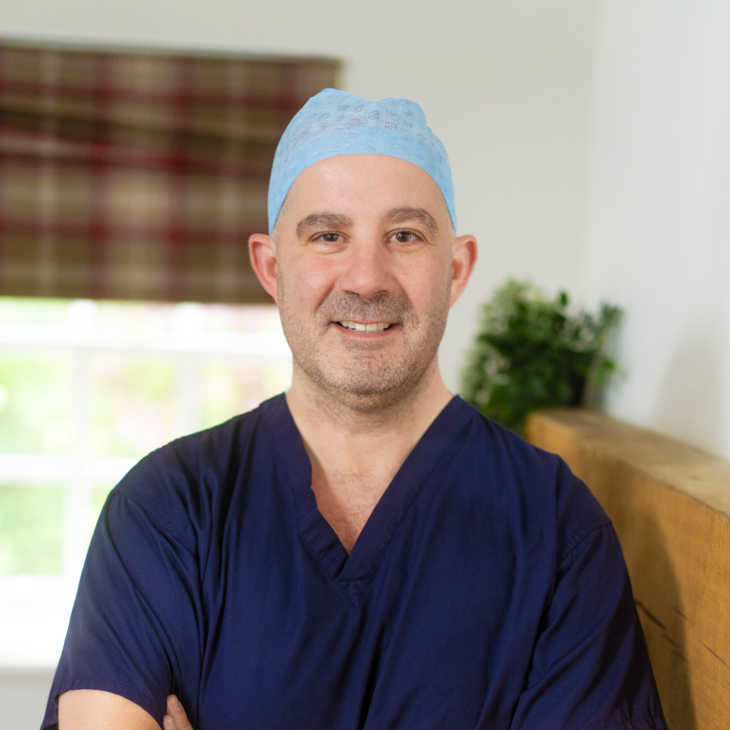 A male healthcare worker wearing blue scrubs and a light blue surgical cap, smiling with arms crossed in a bright room with a window and a plant in the background.
