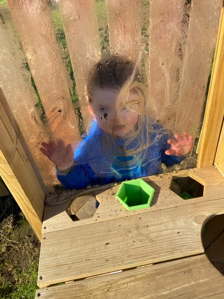A young child pressing their face and hands against a foggy, dirty glass window, inside a wooden playhouse with colorful toys.
