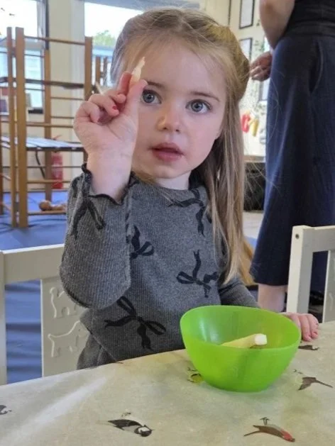 A young girl with long blonde hair and blue eyes holding a small object up near her face at a table with a green bowl in front of her, in a room that appears to be a classroom or play area.