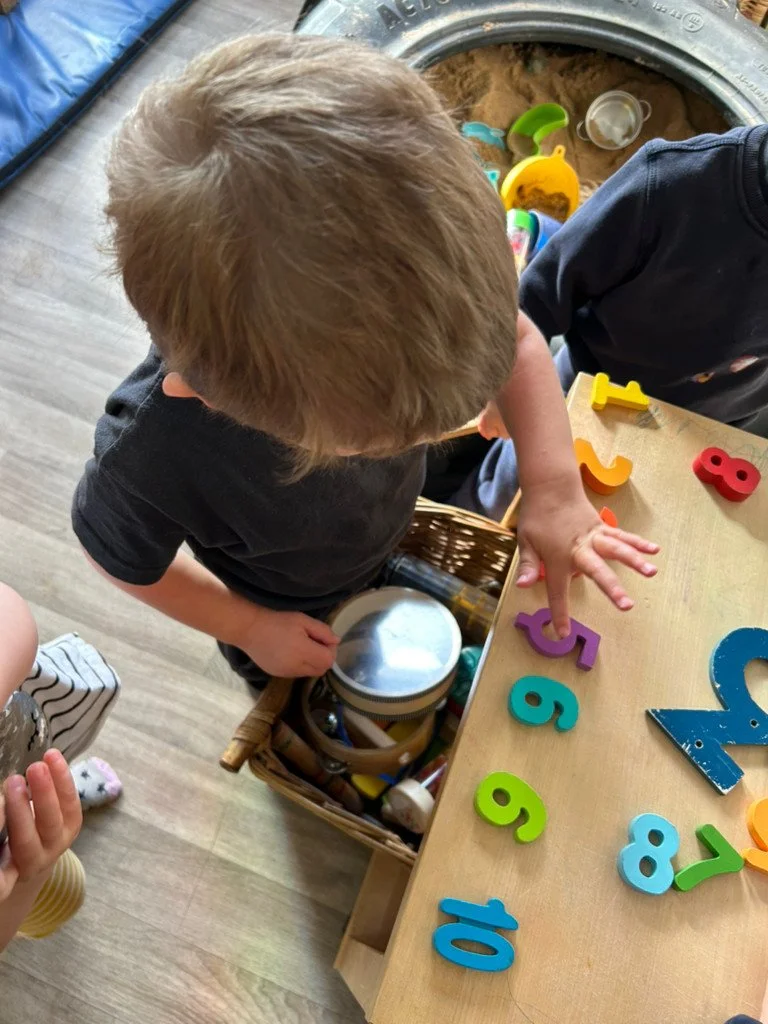 Child playing with colorful foam numbers on a wooden table, with a basket of toys and sandpit in the background.