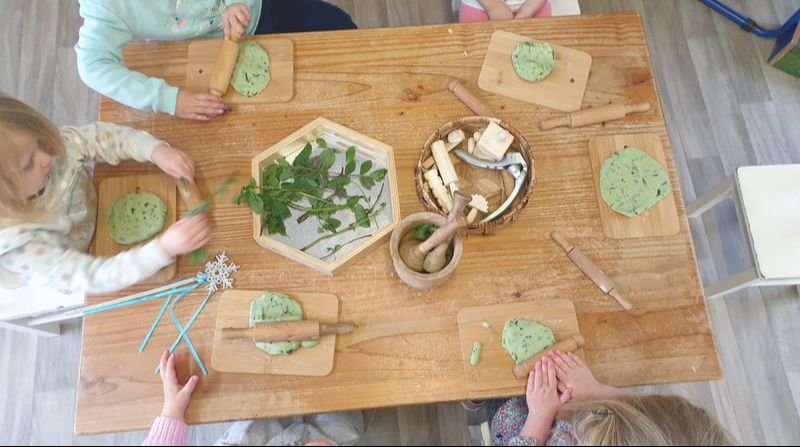 Children sitting around a wooden table, each with a piece of green dough on small wooden boards, using rolling pins to flatten the dough, with natural wooden tools and a plant in the center of the table.