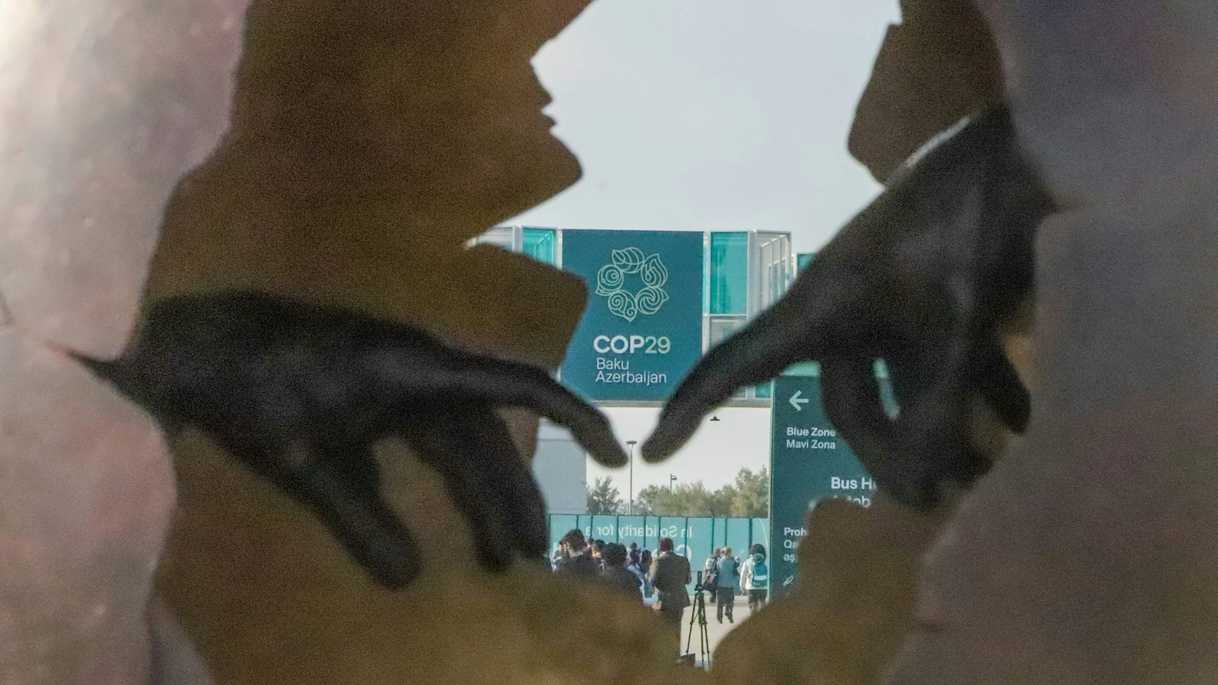 View through a hole in a wall showing a crowd of people, a large sign for COP29 in Baku, Azerbaijan, and a directional sign indicating the Blue Zone and bus stop.