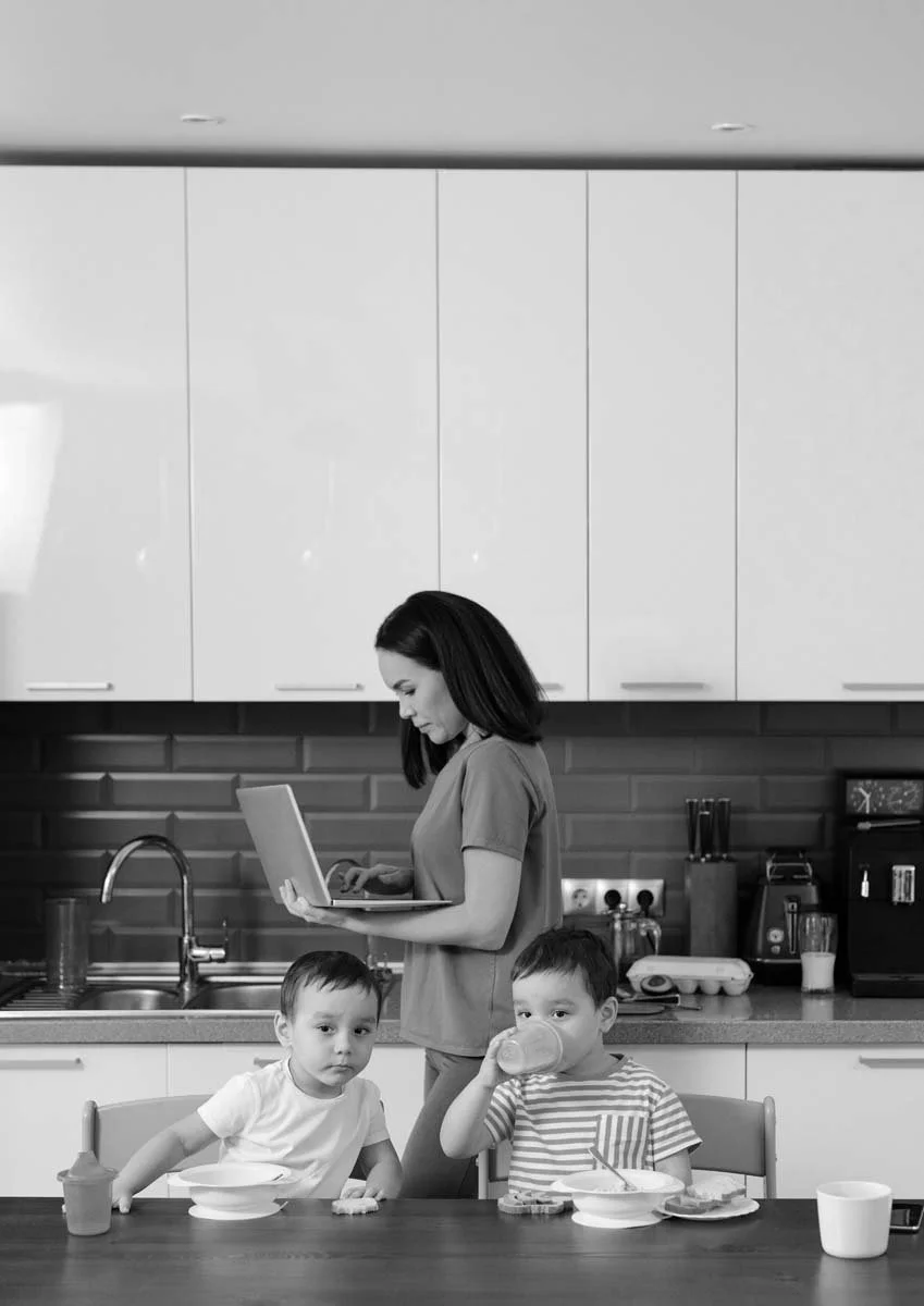 A woman is standing in a kitchen, looking at a laptop. Two young boys are sitting at a table; one is looking at the camera, and the other is drinking from a glass.