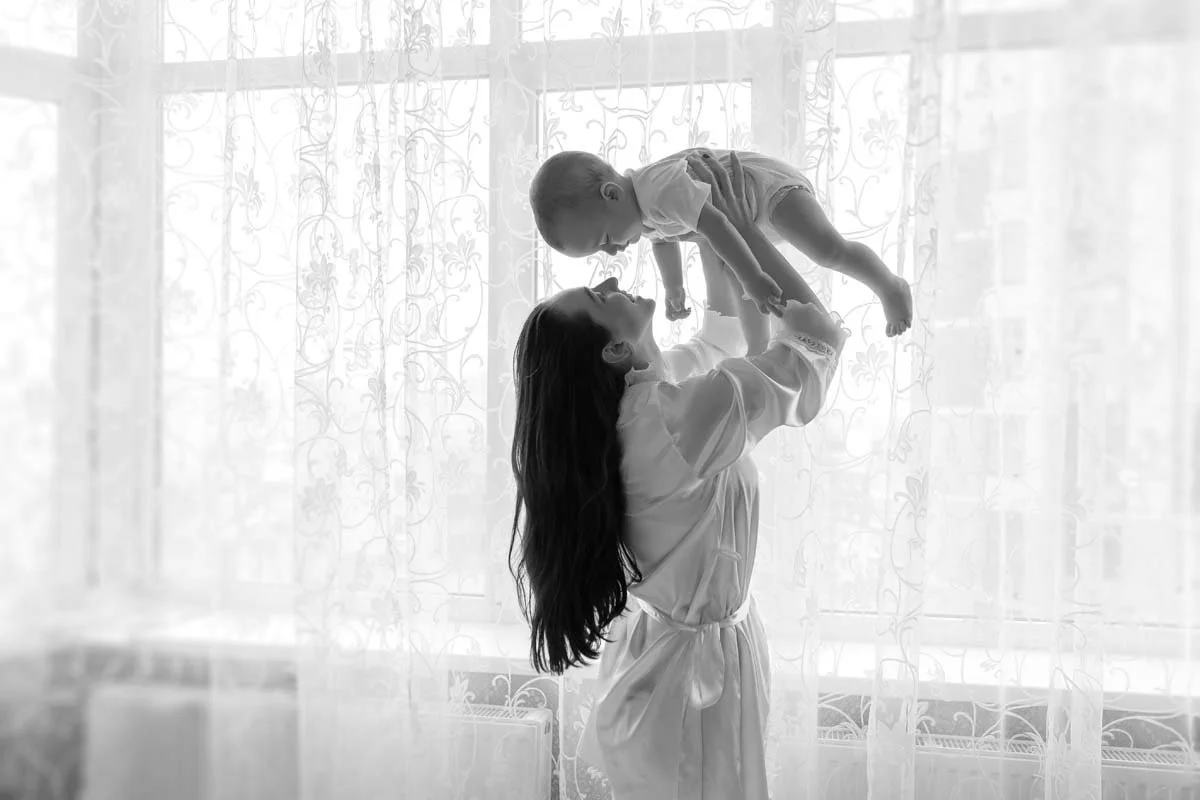 A woman lifting a small child near a window with sheer curtains, both looking at each other, in black and white.