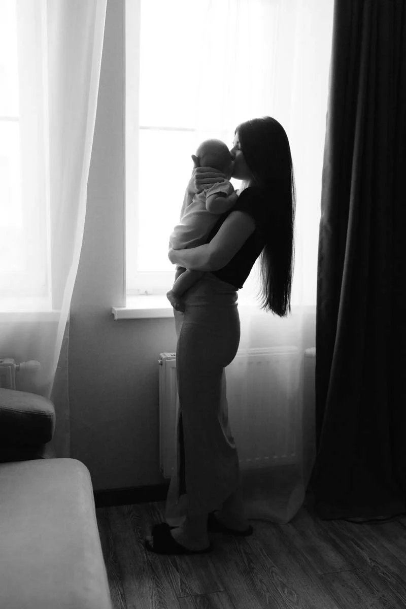A woman holding and gently kissing a baby near a window, silhouette in black and white.