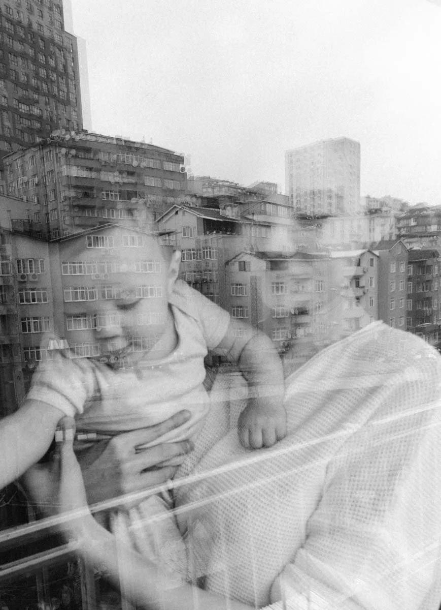 A reflection of a person holding a baby, seen through a window with city buildings in the background.