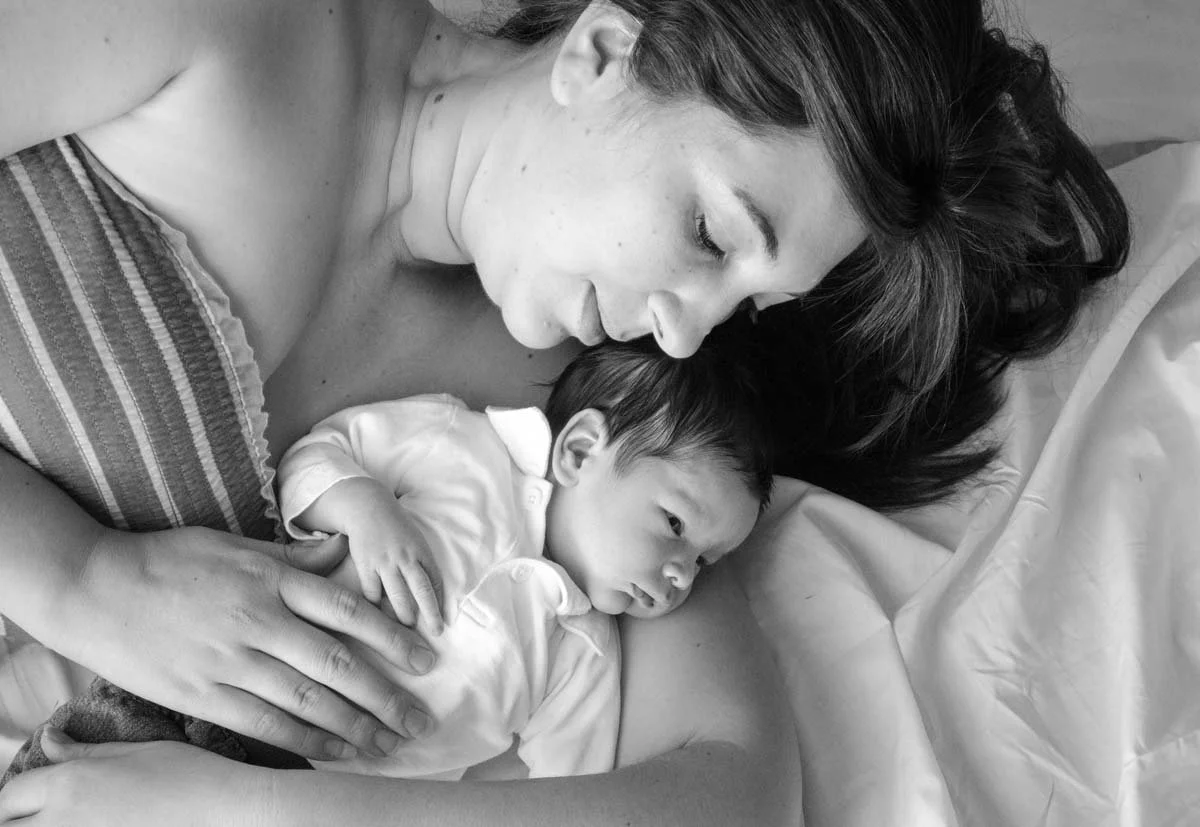 A woman lying in bed, holding a newborn baby close to her chest, both resting peacefully.