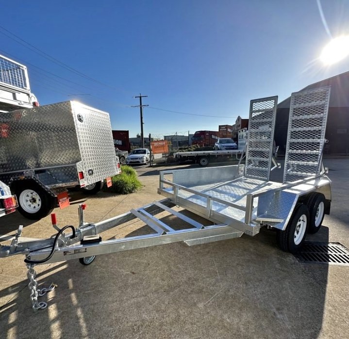 Silver utility trailer with open metal ramp and side rails, parked on concrete in a dealership lot with other trailers and vehicles, under a clear blue sky.