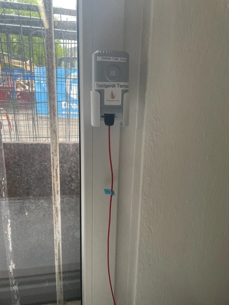 Temperaturmessgerät befestigt an einem Fensterrahmen mit einem roten Kabel, im Hintergrund eine Metallzaun und draußen einige Fahrzeuge und Bäume.