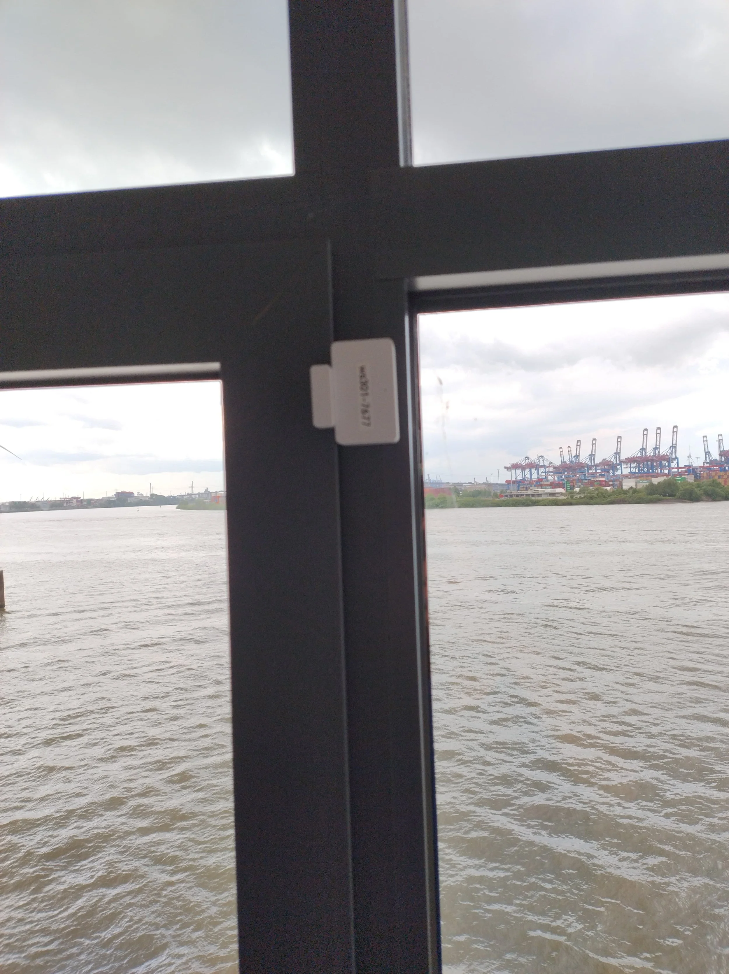 Blick durch eine Fensterscheibe auf einen Fluss mit einem Industriehafen im Hintergrund unter einem bewölkten Himmel.