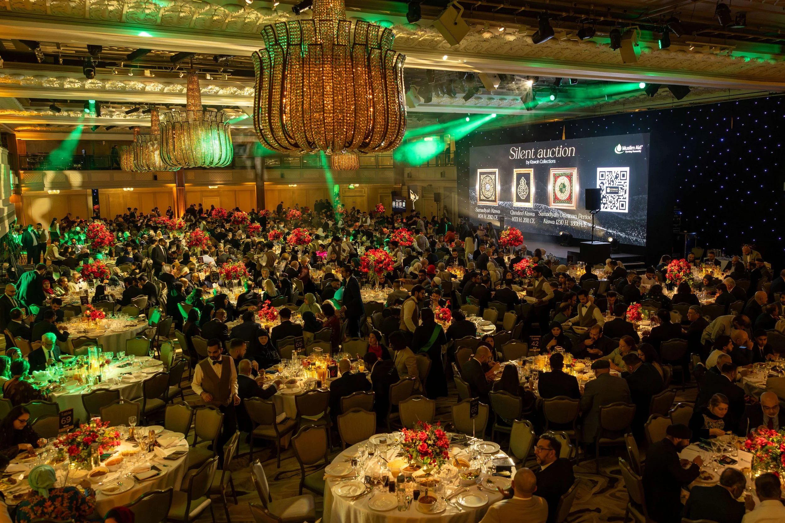 A large banquet hall filled with round tables decorated with pink and red floral centerpieces and covered with white tablecloths, hosting many elegantly dressed guests attending a formal event. The hall features large ornate chandeliers, green lighti