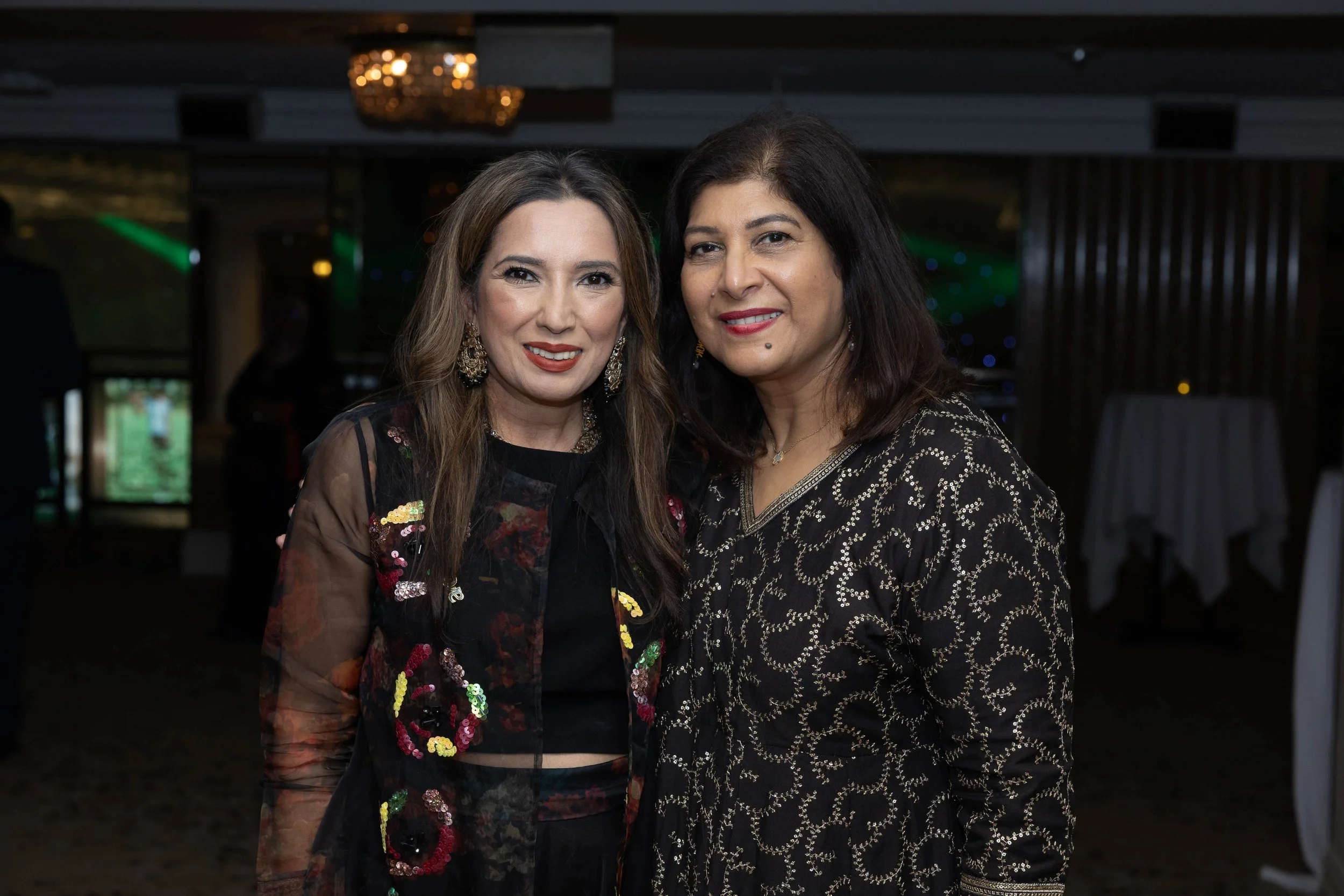 Two women in elegant dresses smiling at a social event or gathering, with a dark background and dim lighting.