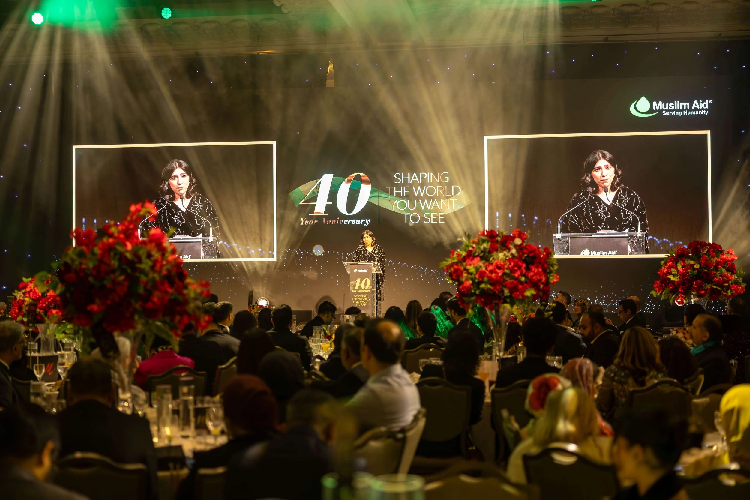 A large indoor event celebrating the 40th anniversary of Muslim Aid, with a woman speaking at a podium on stage, flanked by two large screens showing her image, and decorated with vibrant floral arrangements.
