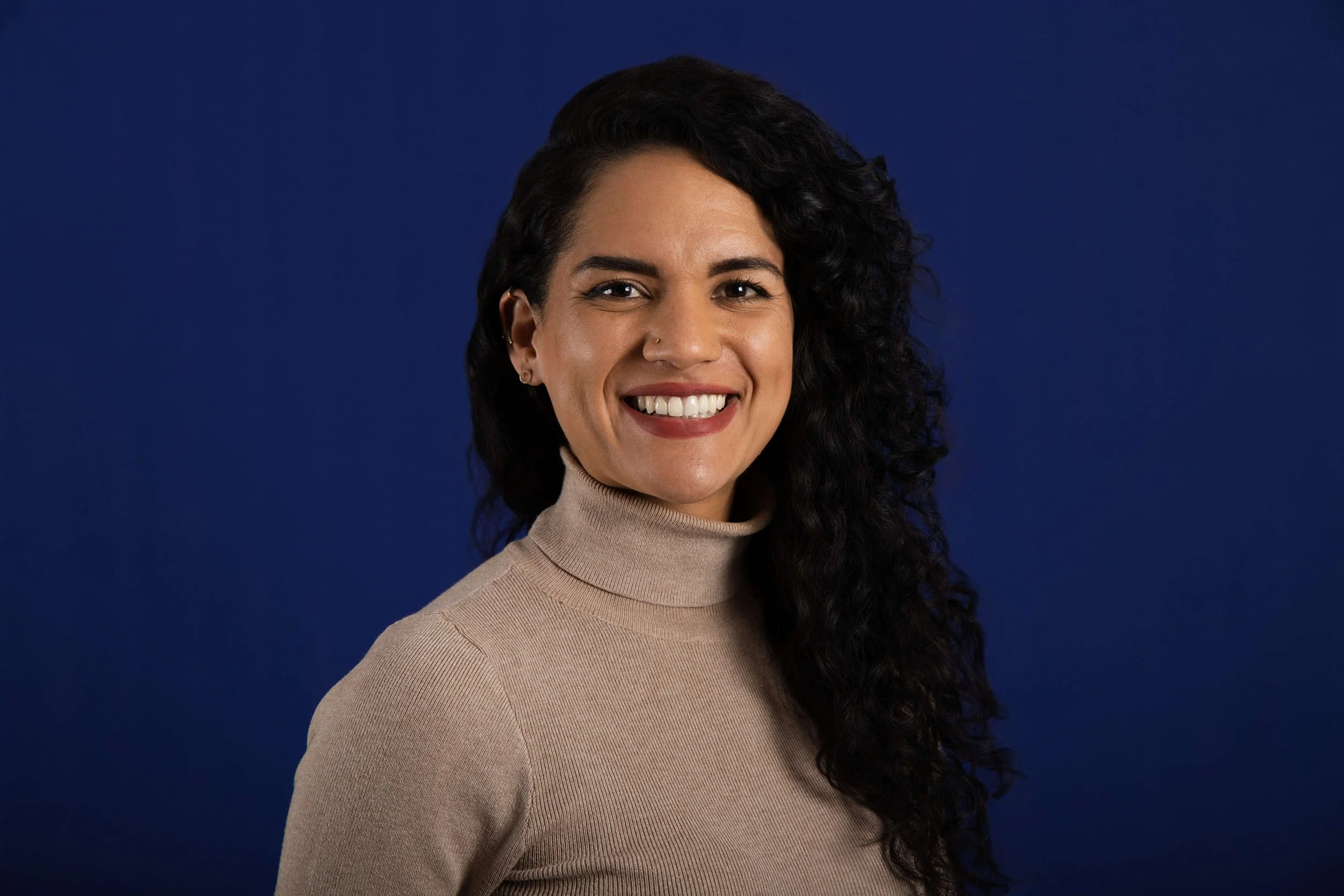 Portrait of a woman with dark curly hair, wearing a beige turtleneck, smiling in front of a dark blue background.