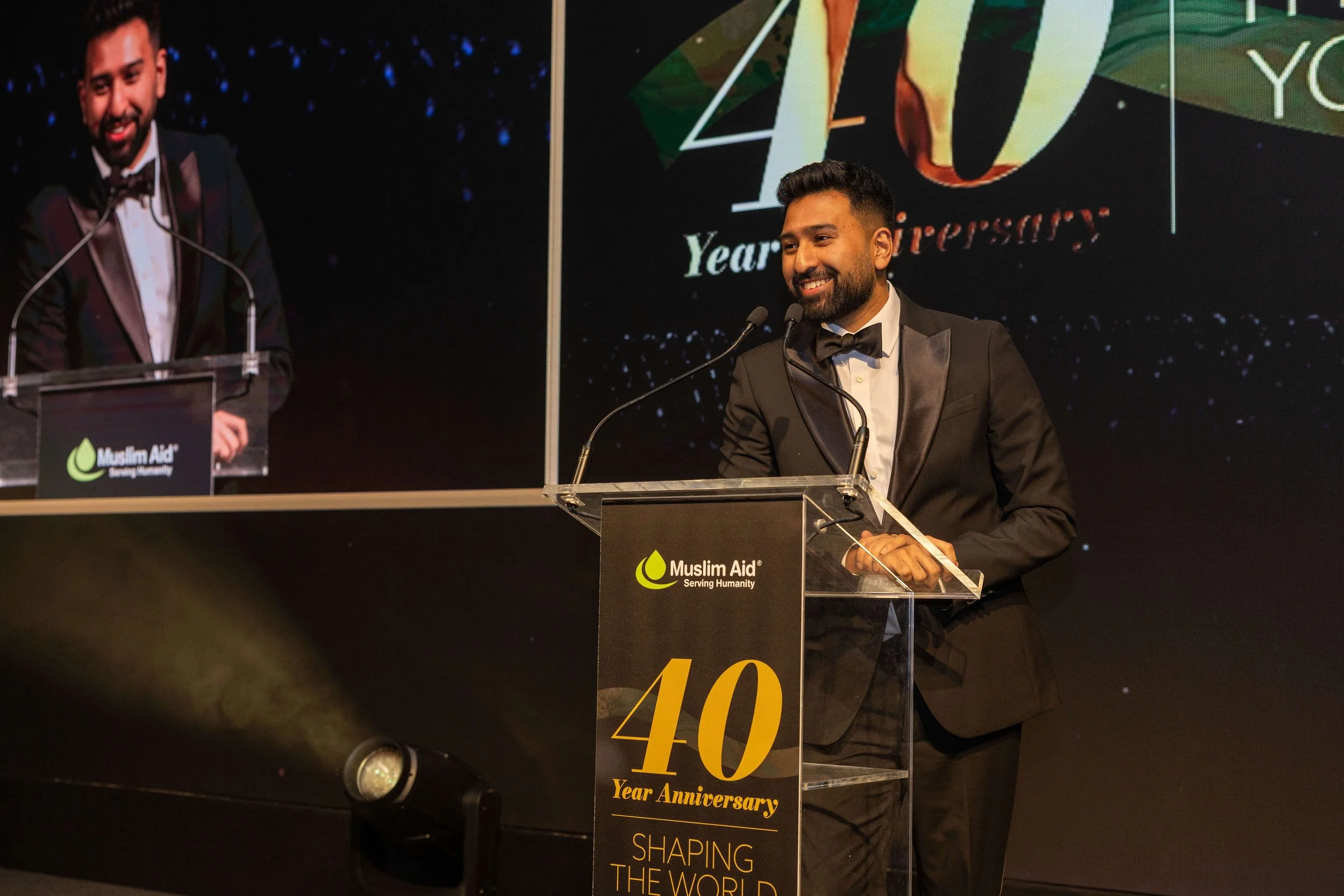 A man in a tuxedo giving a speech at a Muslim Aid event celebrating their 40th anniversary, standing behind a clear podium with a black banner and text, with a large screen in the background displaying the event logo and additional imagery.