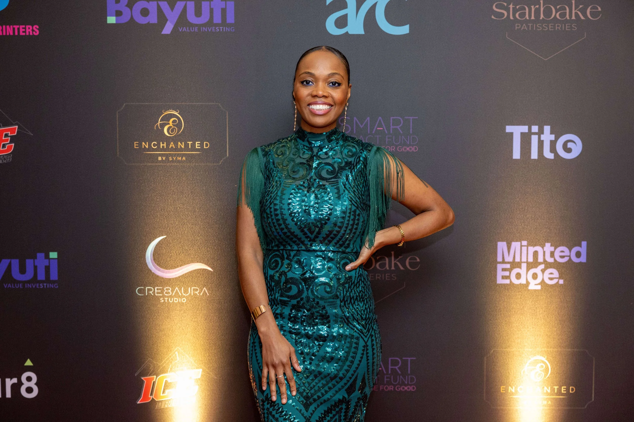 A woman in an elegant teal and black dress with intricate patterns and fringed sleeves, standing and smiling in front of a black backdrop with multiple colorful logos.