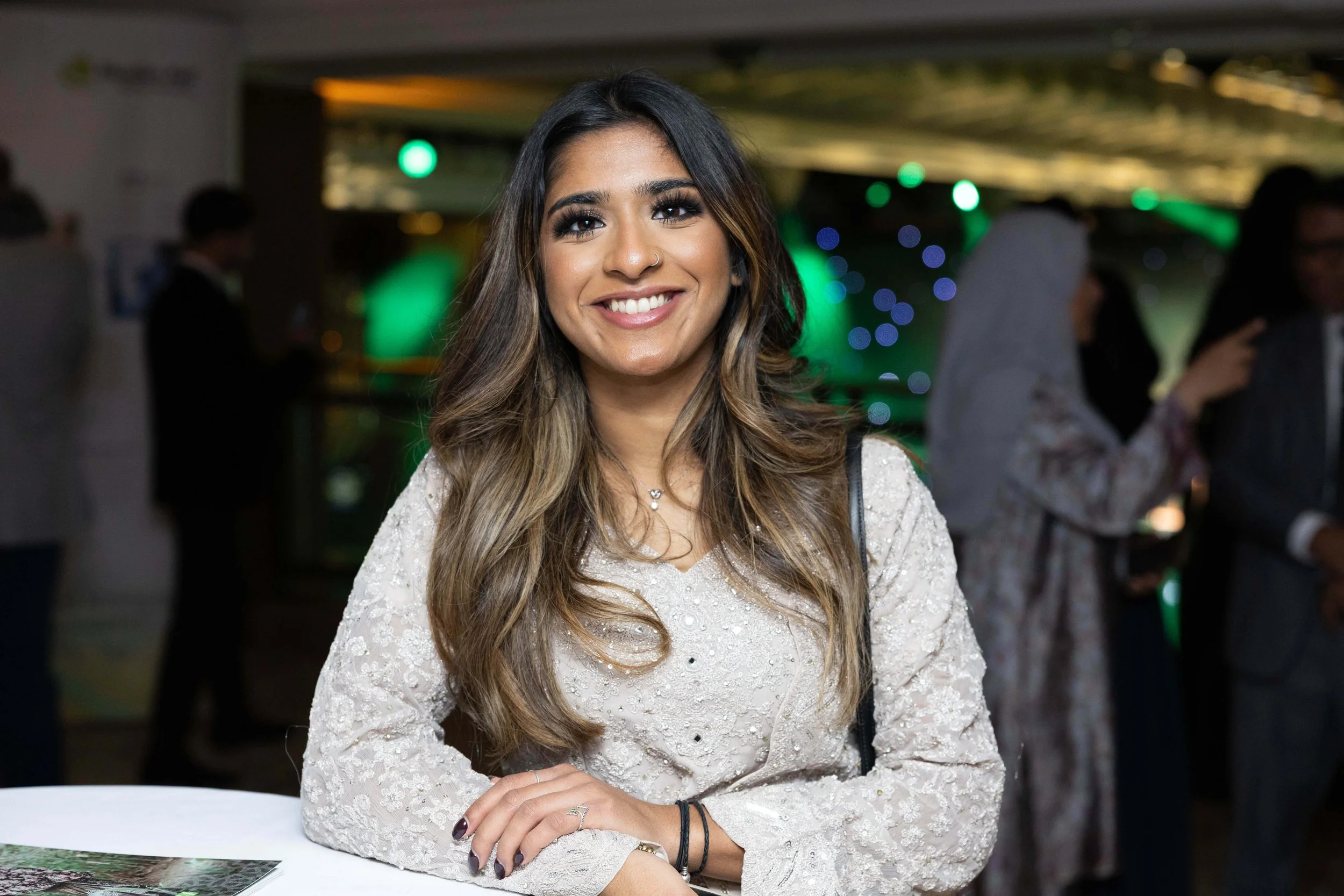 A smiling woman with long wavy hair, wearing a cream-colored lace dress, at a social event indoors with colorful background lights and people talking in the background.